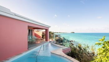 This image features a scenic view of a seaside villa with a pink exterior, an infinity pool, and outdoor seating, overlooking clear blue ocean water.