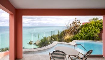 A picturesque view of a coastal scene featuring an infinity pool, a patio with a table and chairs, and vibrant pink walls setup on a terrace.
