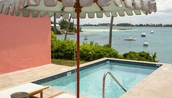 A sunny poolside scene with lounge chairs, a striped umbrella, and a view of the ocean and boats beyond.