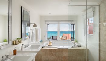 The image shows a luxurious bathroom with double sinks, a glass-enclosed shower, and a bathtub, overlooking a serene ocean view through a large window.