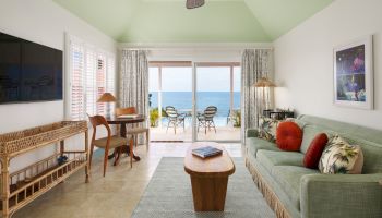 A living room with beach-themed decor, green sofa, wooden furniture, wall art, TV, and glass doors leading to a patio with an ocean view ending the sentence.