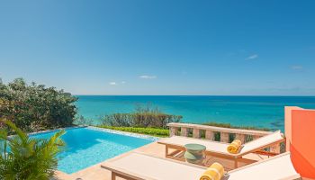 A beautiful seaside pool with two lounge chairs topped with rolled yellow towels overlooks a pristine, turquoise ocean under a clear blue sky.