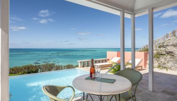 A beautiful seaside patio with an ocean view, featuring a table, chairs, an infinity pool, and a bottle of wine.