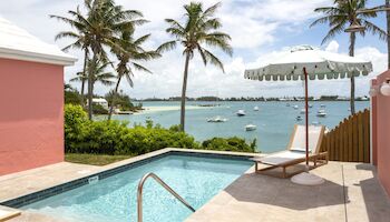 A sunny seaside pool area with turquoise water, palm trees, lounge chairs, and a shaded umbrella overlooking a calm bay with boats, paradise vibes.