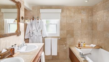 A bathroom with a double sink, mirror, bathtub, and shower. Two robes, towels, and toiletries are visible, with beige tile walls and flooring.