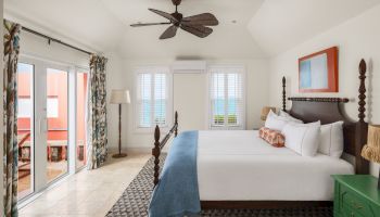 A bright, airy bedroom with a large bed, ceiling fan, balcony, and beach-themed decor, opening to a patio with ocean views.