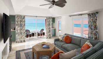 The image shows a bright living room with ocean views, featuring a cozy seating area, a TV, a ceiling fan, and sliding glass doors leading to a balcony.