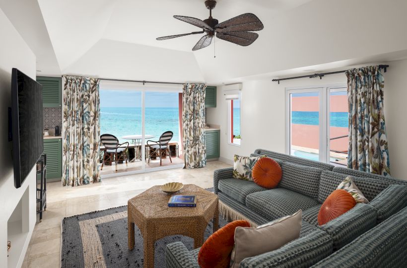 The image shows a bright living room with ocean views, featuring a cozy seating area, a TV, a ceiling fan, and sliding glass doors leading to a balcony.