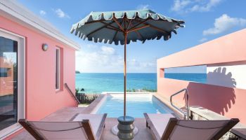 The image shows a relaxing poolside scene with two lounge chairs under a large umbrella, overlooked by a pink building, and with a stunning ocean view.