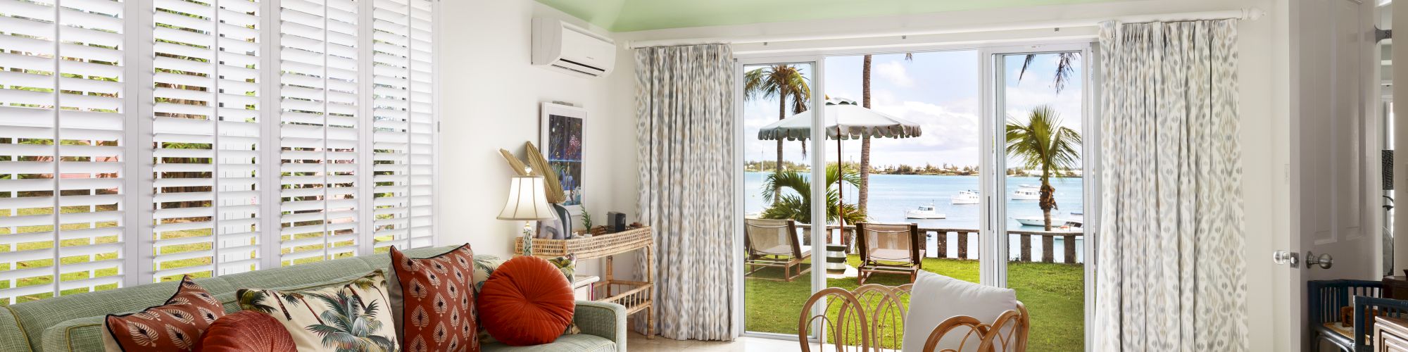 A stylish living room features a green couch with red pillows, wooden furniture, a ceiling fan, and large windows opening to a scenic outdoor view.