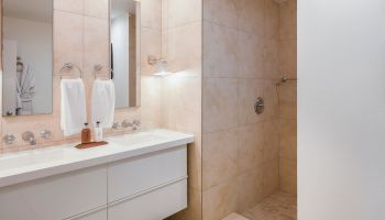 A modern bathroom with a double sink vanity, large mirrors, towel racks, a walk-in shower, and beige tiles on the walls and floor.