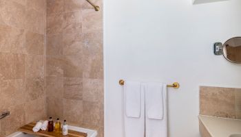 The image shows a bathroom with a beige-tiled bathtub, two white towels on a golden towel bar, and toiletries on a wooden tray in the tub.