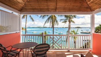 A balcony with a table and chairs overlooks a scenic view of a beach with palm trees and boats on the water, under a partly cloudy sky.