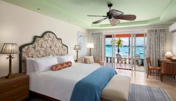 A well-decorated, tropical-themed bedroom with a king-sized bed, ceiling fan, and a balcony overlooking the ocean. The room has wooden furniture.