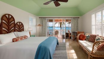 A well-decorated bedroom with a large bed, blue blanket, seating area, ceiling fan, and a view of the ocean through glass doors ending the sentence.