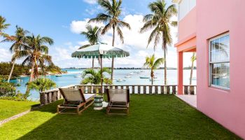 A tropical setting with palm trees, a pink building, two lounge chairs, and an umbrella by a waterfront, all under a clear blue sky with scattered clouds.