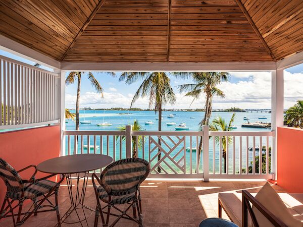 A covered balcony with a wooden ceiling overlooks a beautiful seaside view featuring palm trees, boats on the water, and a clear, sunny sky.