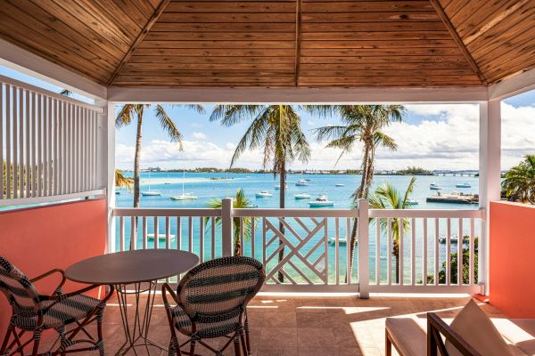 A covered balcony with a wooden ceiling overlooks a beautiful seaside view featuring palm trees, boats on the water, and a clear, sunny sky.