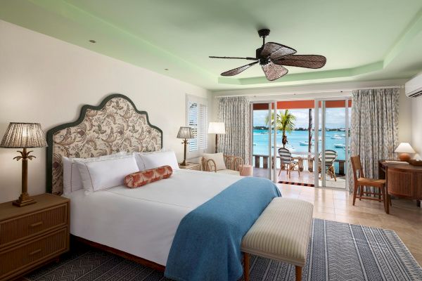 The image shows a spacious bedroom with a large bed, tropical-style decor, and a balcony with outdoor seating that overlooks the ocean.