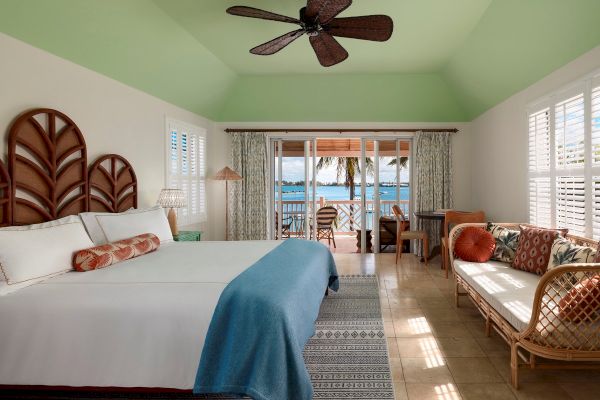 This image shows a bright and airy bedroom with a large bed, a ceiling fan, a couch, and a window with a view of the ocean and palm trees outside.