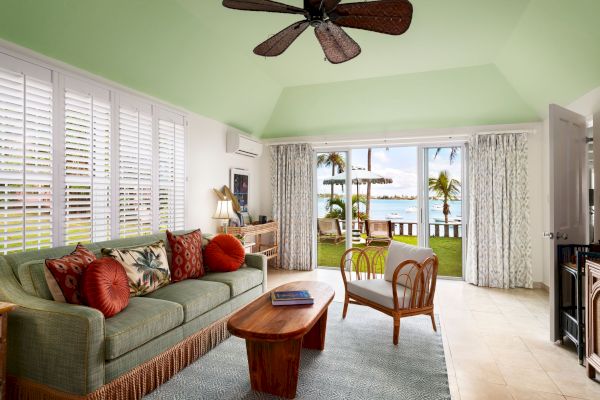 A modern tropical living room with a ceiling fan, green sofa, wooden furniture, and large windows opening to a patio with a beach view ending the sentence.