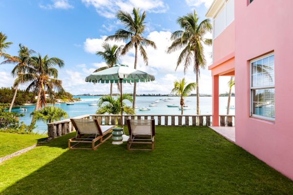 A seaside scene with two lounge chairs under a striped umbrella, surrounded by palm trees, a pink building, and a view of boats and the ocean.