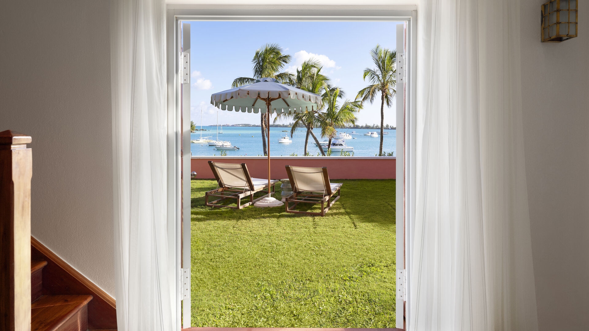 Image shows an open doorway leading to a sunny yard with lounge chairs under an umbrella, palm trees, and a view of the ocean with boats in the distance.