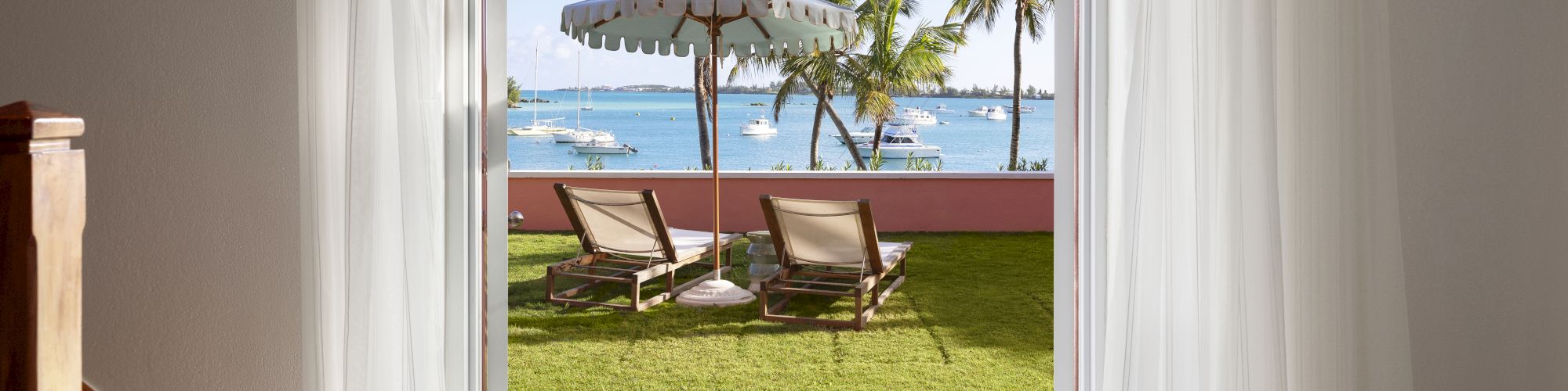 Image shows an open doorway leading to a sunny yard with lounge chairs under an umbrella, palm trees, and a view of the ocean with boats in the distance.