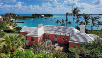 A tropical coastal scene with a pink house surrounded by lush greenery, overlooking a blue sea under a partly cloudy sky dotted with palm trees.