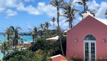 A picturesque scene with a pink house, palm trees, lush greenery, and a view of a turquoise ocean under a clear blue sky, ending the sentence.