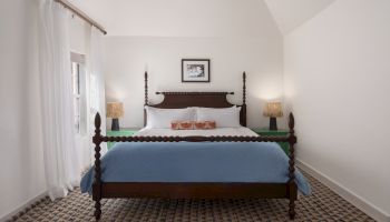 A neatly arranged bedroom features a wooden four-poster bed with a blue blanket, two bedside tables with lamps, white walls, and a large window.