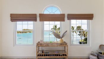 A room with three large windows showing a beach view, a wicker table with decorative items, and a patterned rug.
