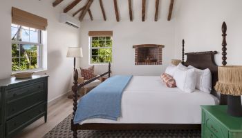A cozy bedroom with wooden beamed ceiling, large bed, green dresser, two windows, wall-mounted AC unit, lamp, and brick-inset on the wall.