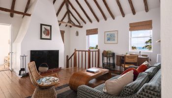 A cozy living room with wooden beams, a fireplace, woven chairs, a sofa, a rustic coffee table, and a desk by the window.
