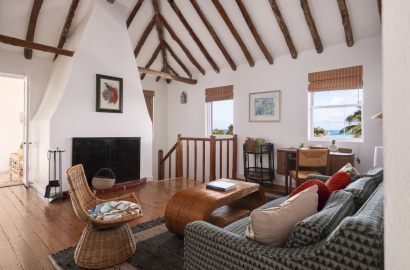 A cozy living room with wooden beams, a fireplace, woven chairs, a sofa, a rustic coffee table, and a desk by the window.
