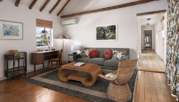 A cozy living room with a slanted wooden-beam ceiling, featuring a sofa, coffee table, wicker chair, desk, and artwork on the walls, with wood flooring.