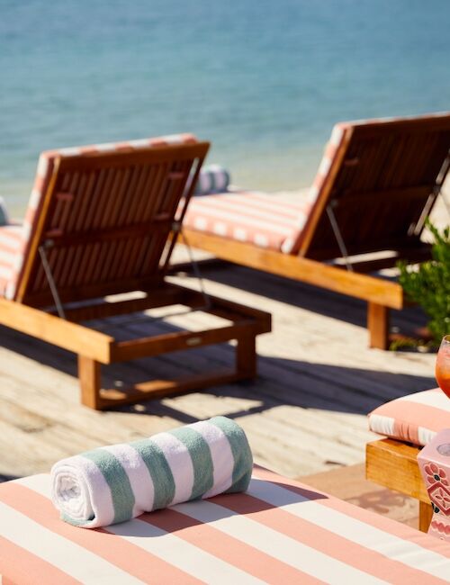 Two poolside sun loungers with striped cushions, towels, and a table with drinks on a sunny deck by the water.