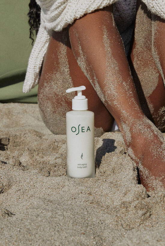 A bottle of OEA lotion stands in sand between sandy feet wrapped in a towel, at the beach, with clear water and sun.