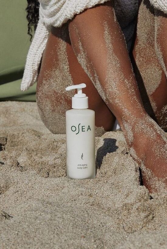 A white pump bottle on sandy beach, labeled &ldquo;Osea,&rdquo; between sun-kissed legs with a towel in the background.