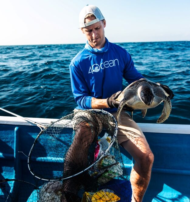 Man on a blue boat holds a large fish he just caught; the ocean stretches blue in the background under a clear sky.