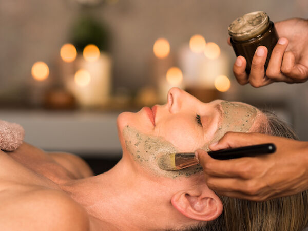 A person receives a facial treatment: a mask is applied with a brush while another hand holds a jar, in a spa setting with soft candlelight.