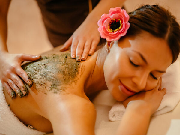 A woman receives a back treatment with a green mud or clay mask, lying face down with a flower in her hair and a relaxing spa setting. End.