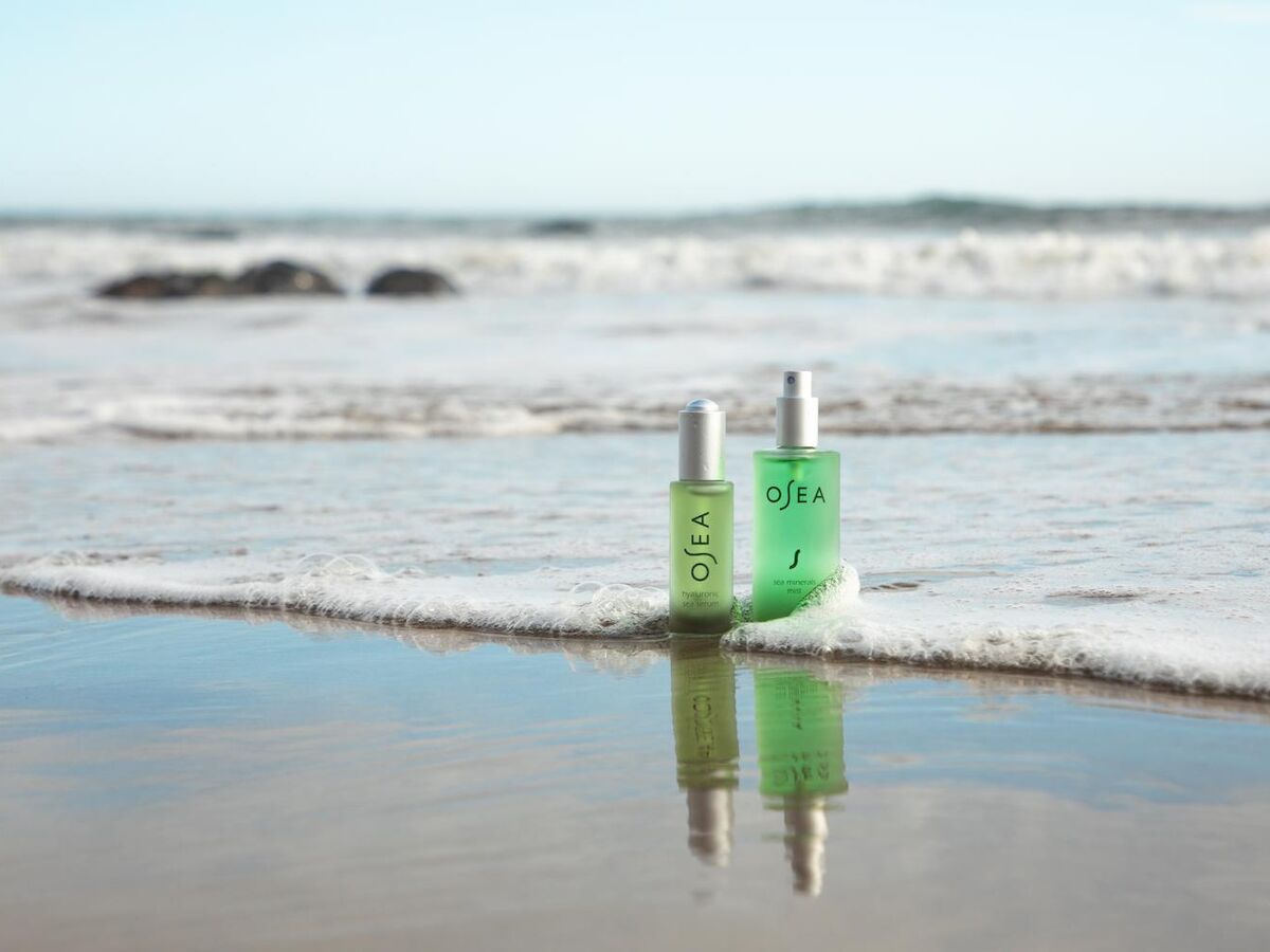 Two green OSEA bottles stand at the water&rsquo;s edge on a sandy beach, their reflections shimmering in a shallow tide pool.