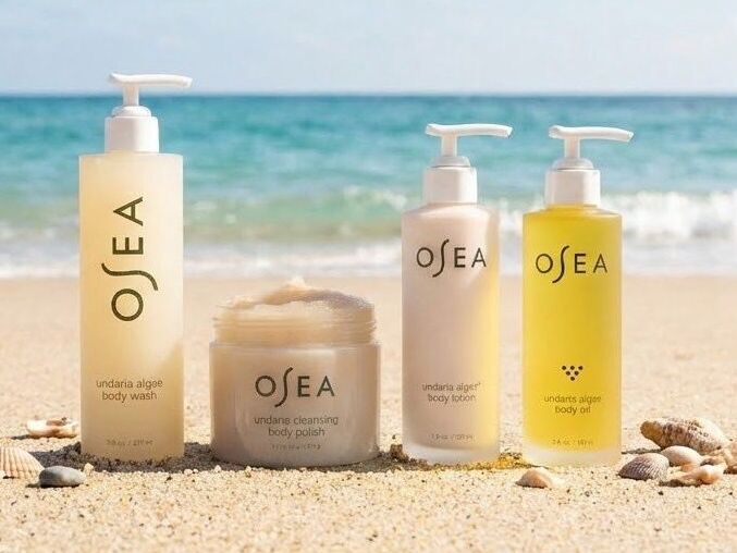 A row of OSEA skincare bottles on a sandy beach with the sea in the background, sunny and bright, product lineup on the shore.