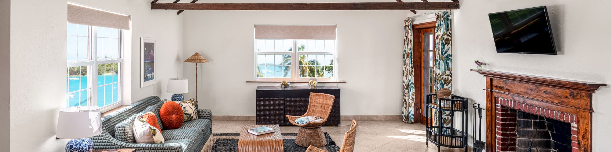 A cozy living room with wooden beams, fireplace, TV, wicker furniture, and large windows offering a scenic view, decorated with modern and rustic touches.