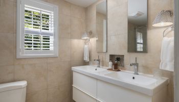 A modern bathroom with beige tiles, a double sink with mirrors, a window with shutters, and a toilet.