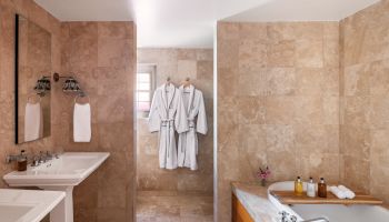The image shows a bathroom with two sinks, towels, a bathtub, and bathrobes hanging on the wall, all surrounded by beige tile walls.