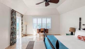 A bright bedroom features a bed with a blue blanket, ceiling fan, desk by a window with shutters, and a door with curtains leading outside.