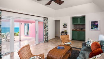 A cozy, tropical-themed living room with wicker furniture, a ceiling fan, a kitchenette, and sliding doors that open to a patio with an ocean view.