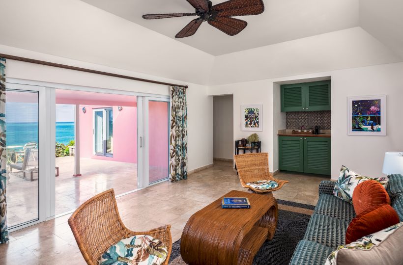 A cozy living room overlooking a patio with a beach view. Includes wicker chairs, a stylish coffee table, a sofa, and a ceiling fan.
